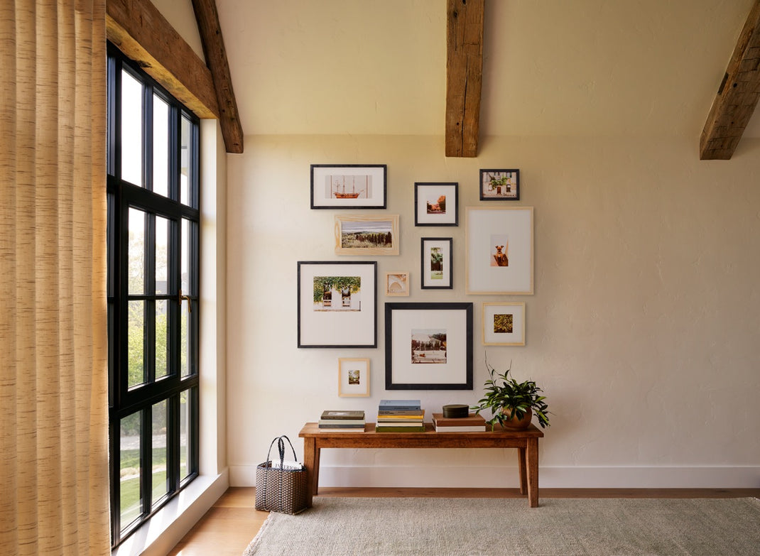A styled interior room corner with a gallery wall of framed art above a wooden bench, next to a large black-framed window with woven curtains.