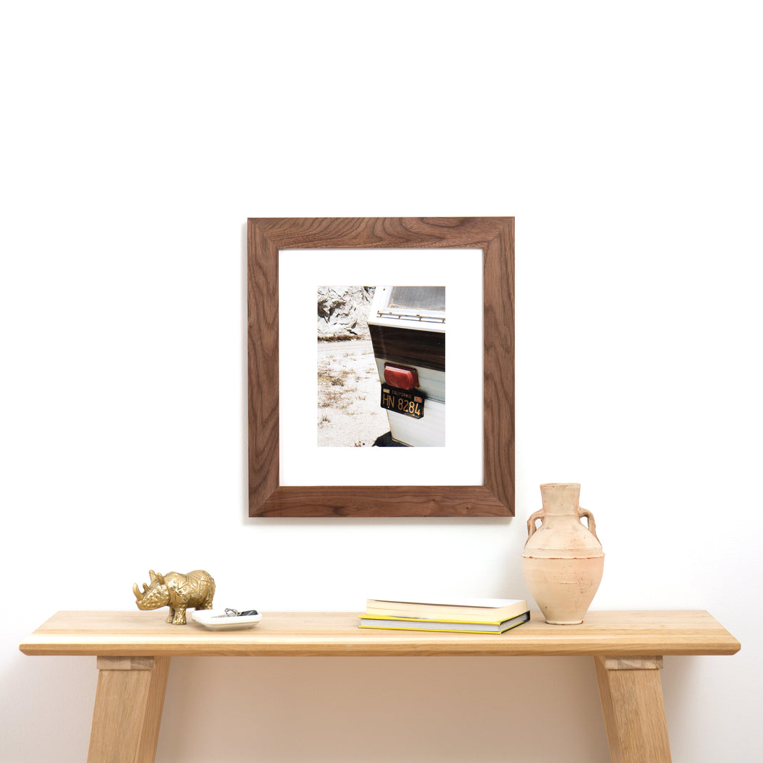 photograph of the license plate on the back of a camper framed in Walnut Wide hardwood frame style with white mat hanging on a white wall above a wood console table