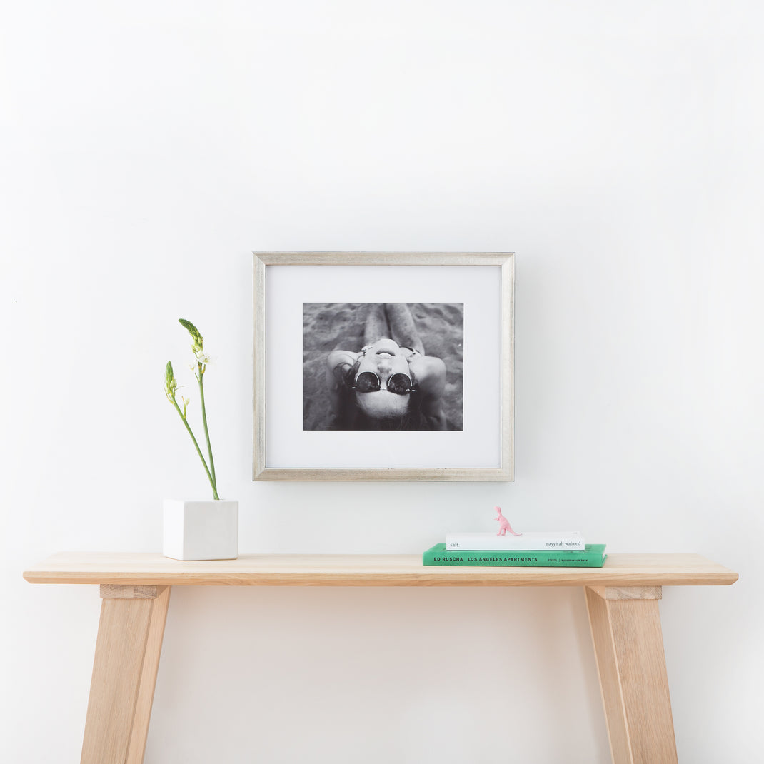 Black & white photo of a woman wearing sunglasses on the beach framed in a silver Beverly frame with white mat hanging on a white wall above a light wood entry table