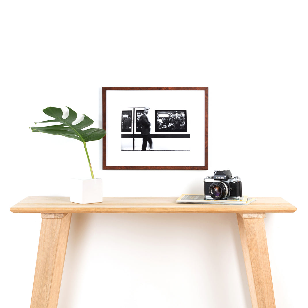 blurry black and white photo of a man in front of a subway train framed in dark skinny burl wood Frederick frame with a white mat hanging on a white wall above a wood console table