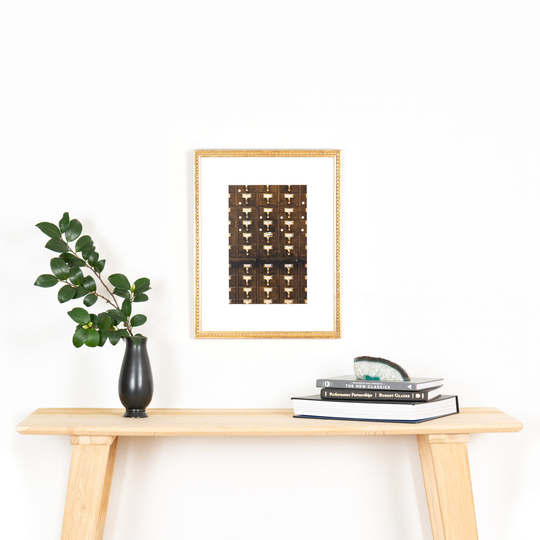 photograph of library drawers framed in tuliped gold Montclare frame with a white mat hanging on a white wall above a wood console table