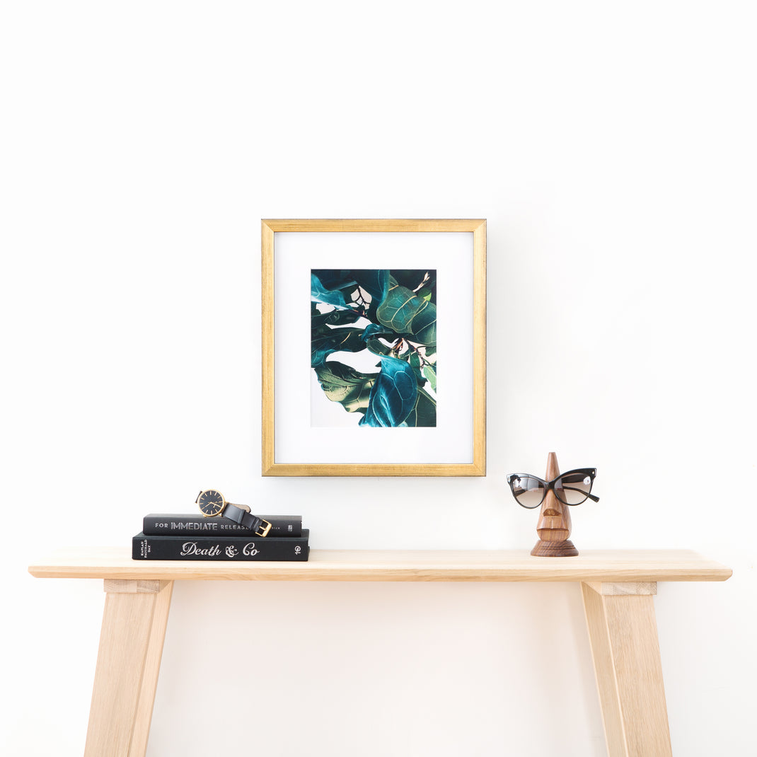 close up photo of fiddle leaf fig leaves framed in gold Olympia frame with a white mat hanging on a white wall above a wood console table