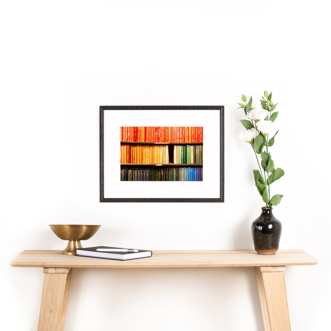 photograph of a bookshelf arranged in rainbow framed in detailed black frame with a white mat hanging on a white wall above a wood console table