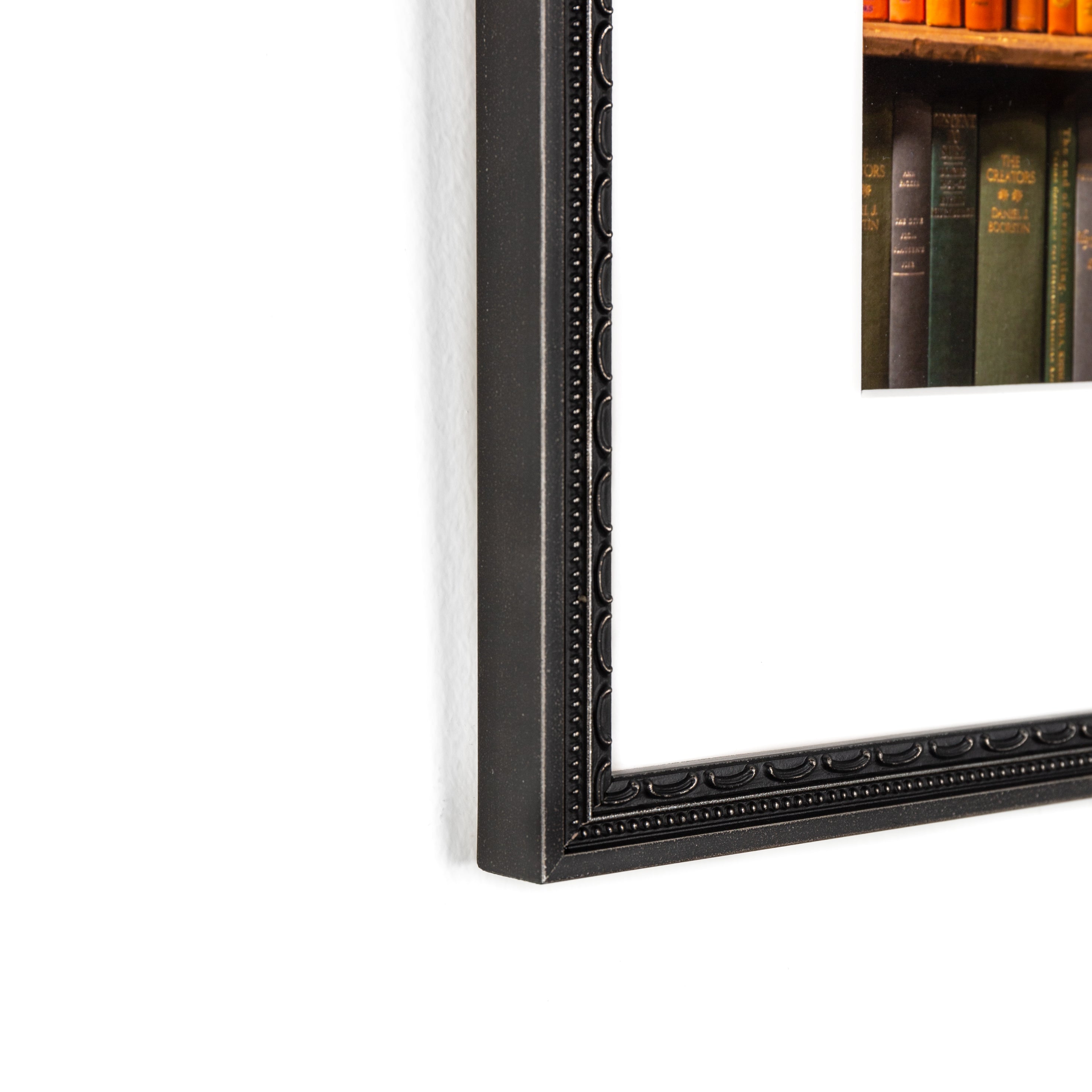 bottom left corner of photograph of a bookshelf arranged in rainbow framed in detailed black frame with a white mat