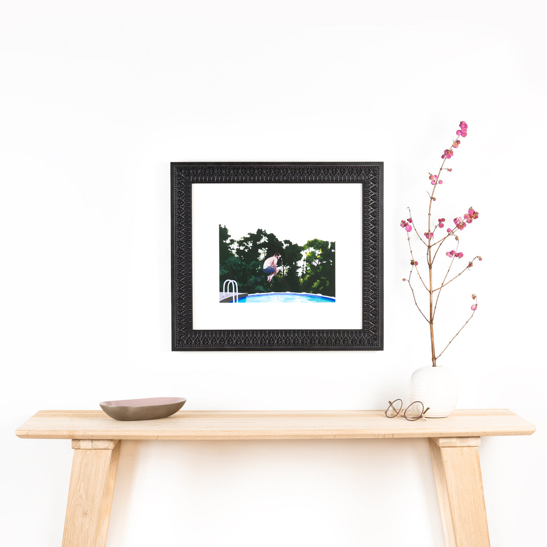 art print of a man doing a cannonball into a pool framed in ornate black Ravenswood frame with a white mat hanging on a white wall above a wooden console table