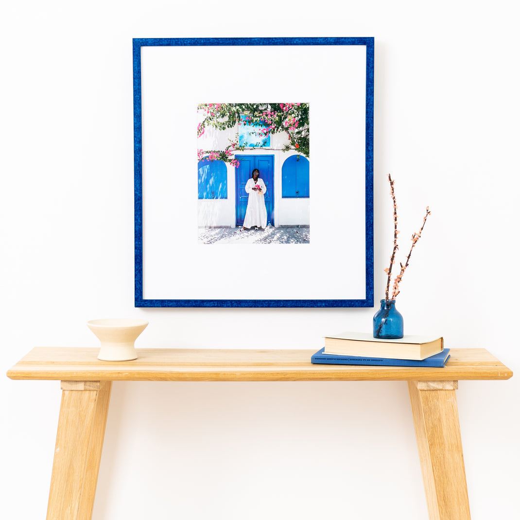 photo of a woman in front of a blue door framed in royal blue glazed Santorini frame with a white mat hanging on a white wall over a wood console table