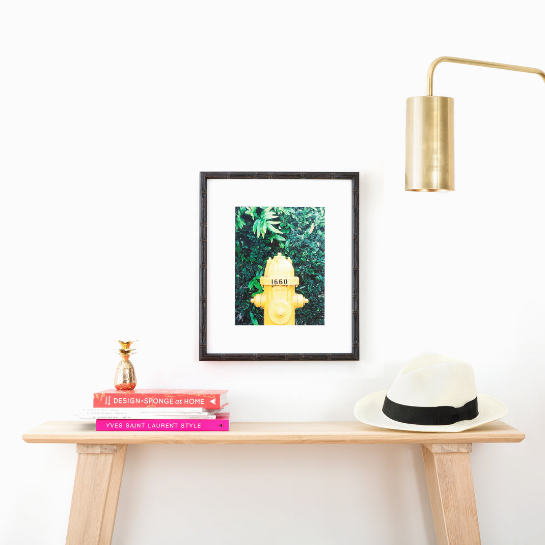 Photo of a yellow fire hydrant framed in a black bamboo Bali frame with a white mat hanging on a white wall above an entry table