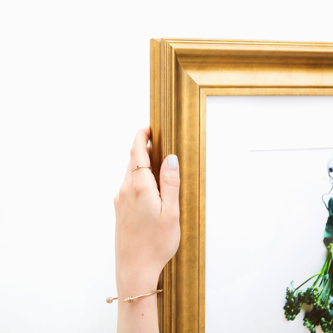 top left corner of a illustration of a woman with a broccolini skirt framed in wide sloped gold Chelsea frame with a white mat