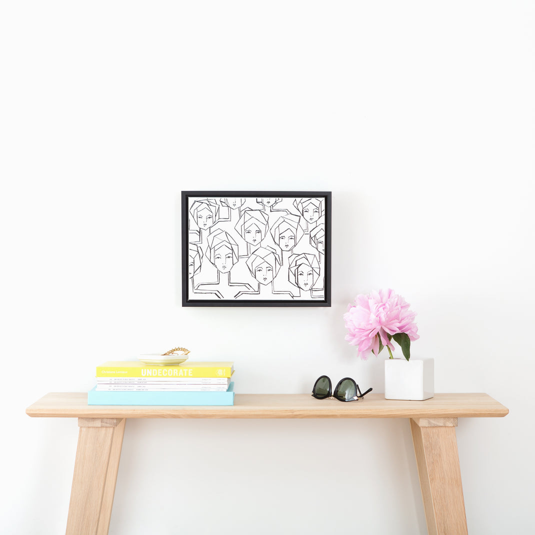 black and white line painting of women on canvas framed in Heathrow black canvas floater frame hanging on a white wall above a wood console table