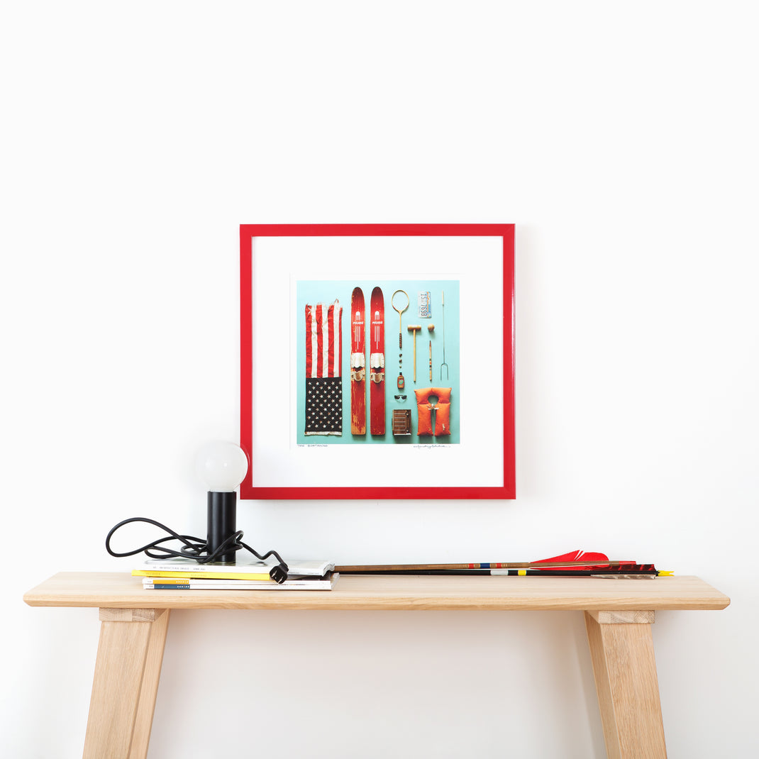 flat lay photograph of outdoor equipment framed in red lacquered Madrid frame with a white mat hanging on a white wall above a wood console table