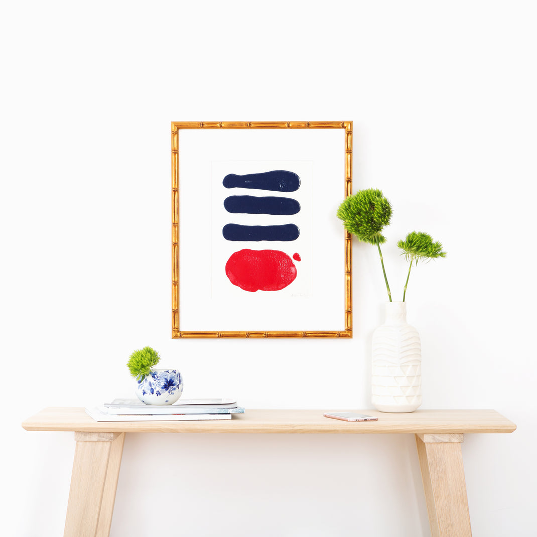 red and blue abstract art framed in Mandalay gold bamboo frame with a white mat hanging on a white wall above a wood console table