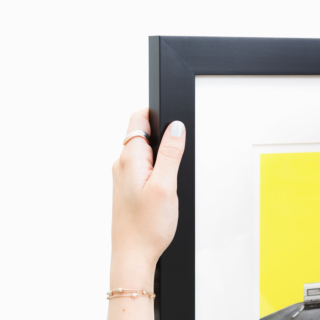 top left corner of abstract print of a camper on a road with yellow sky framed in modern black Mercer frame with a white mat
