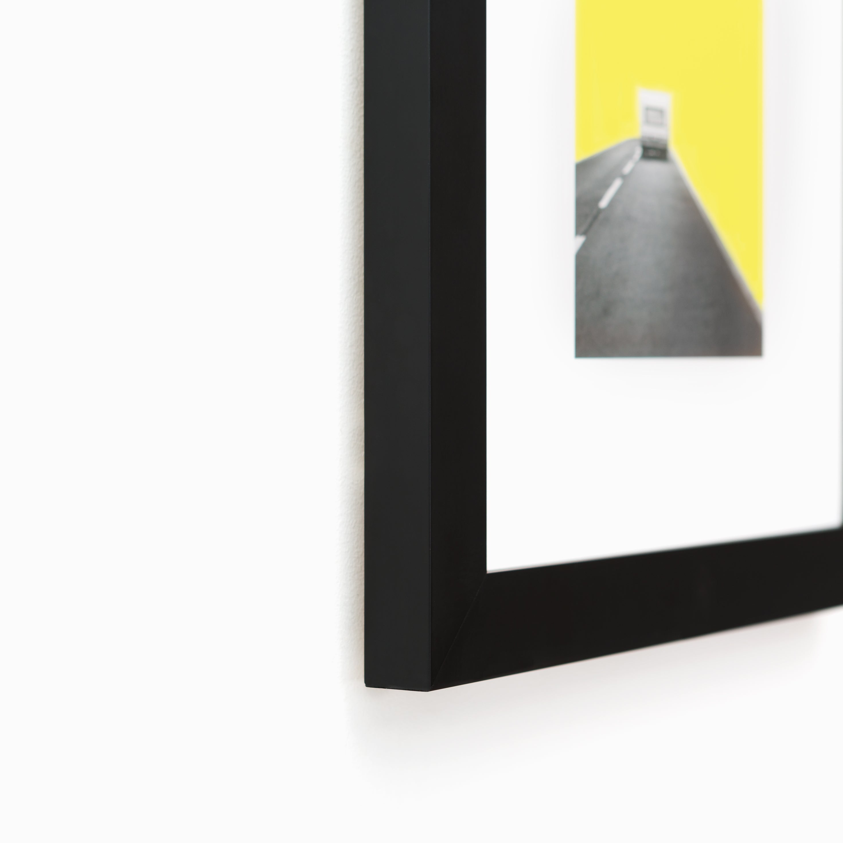 bottom left corner of abstract print of a camper on a road with yellow sky framed in modern black Mercer frame with a white mat