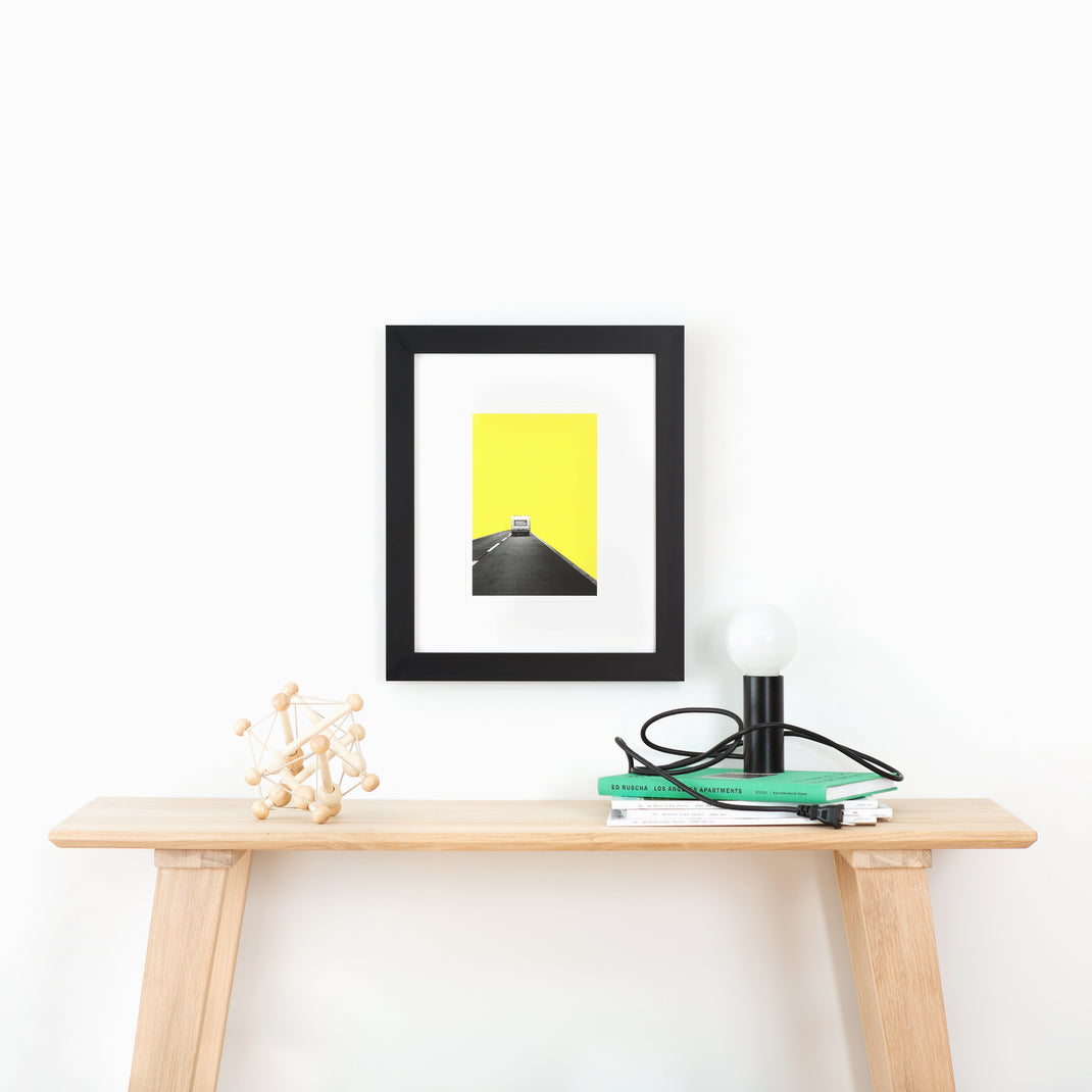 abstract print of a camper on a road with yellow sky framed in modern black Mercer frame with a white mat hanging on a white wall above a wood console table