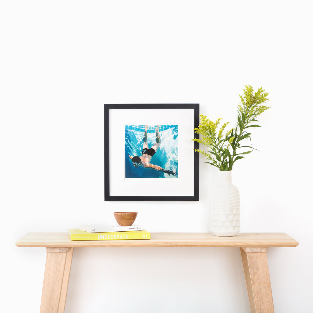 art print of a man swimming in a pool framed in thin black gallery Mercer Slim frame with a white mat hanging on a white wall above a wood console table