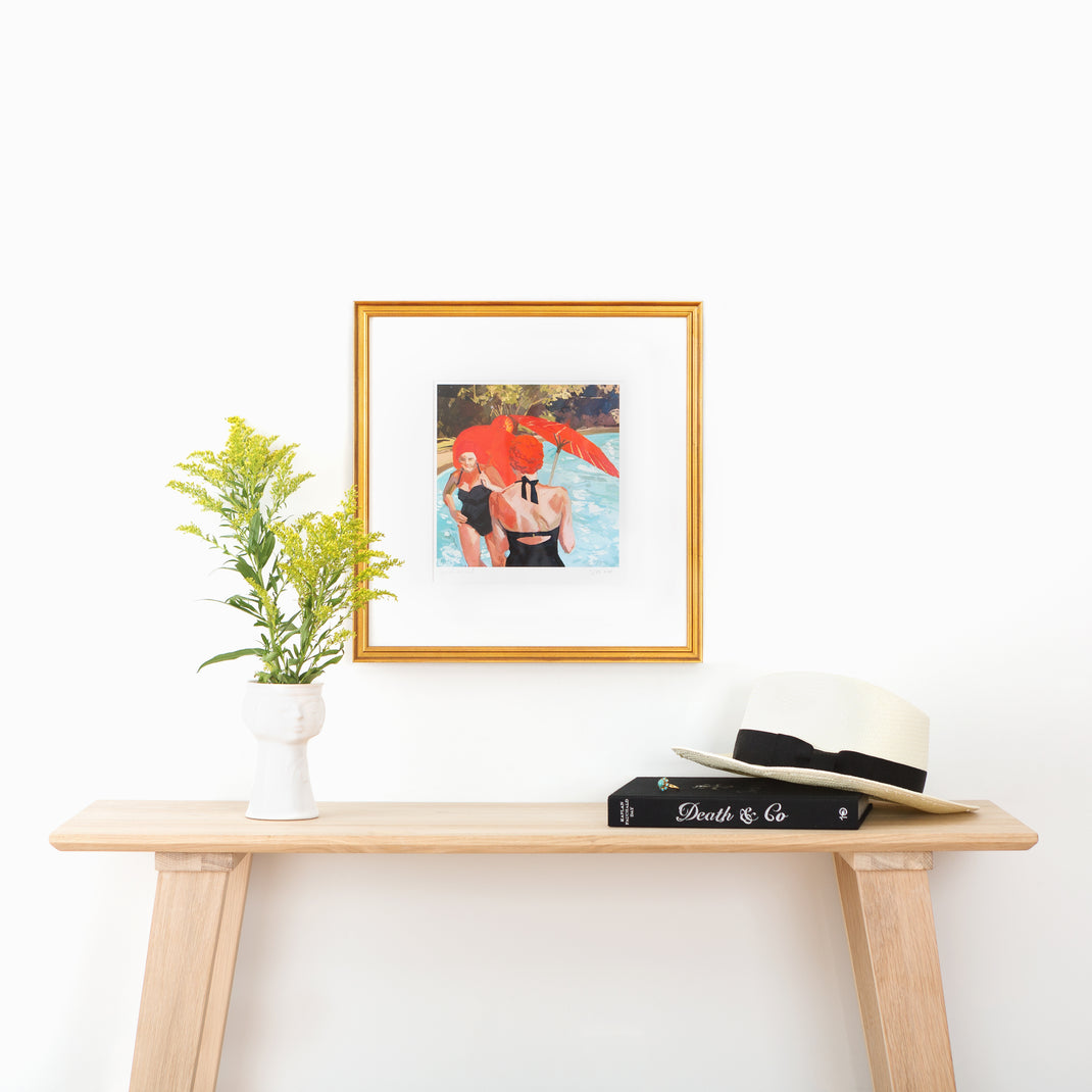 art print of women by a pool in vintage bathing suits framed in Richmond gold frame with a white mat hanging on a white wall above a wood console table