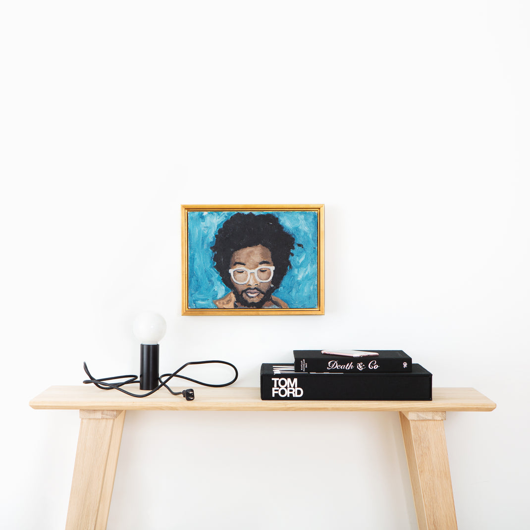 painted portrait of a man wearing white glasses framed in Valencia gold floater style canvas frame hanging on a white wall above a wood console table